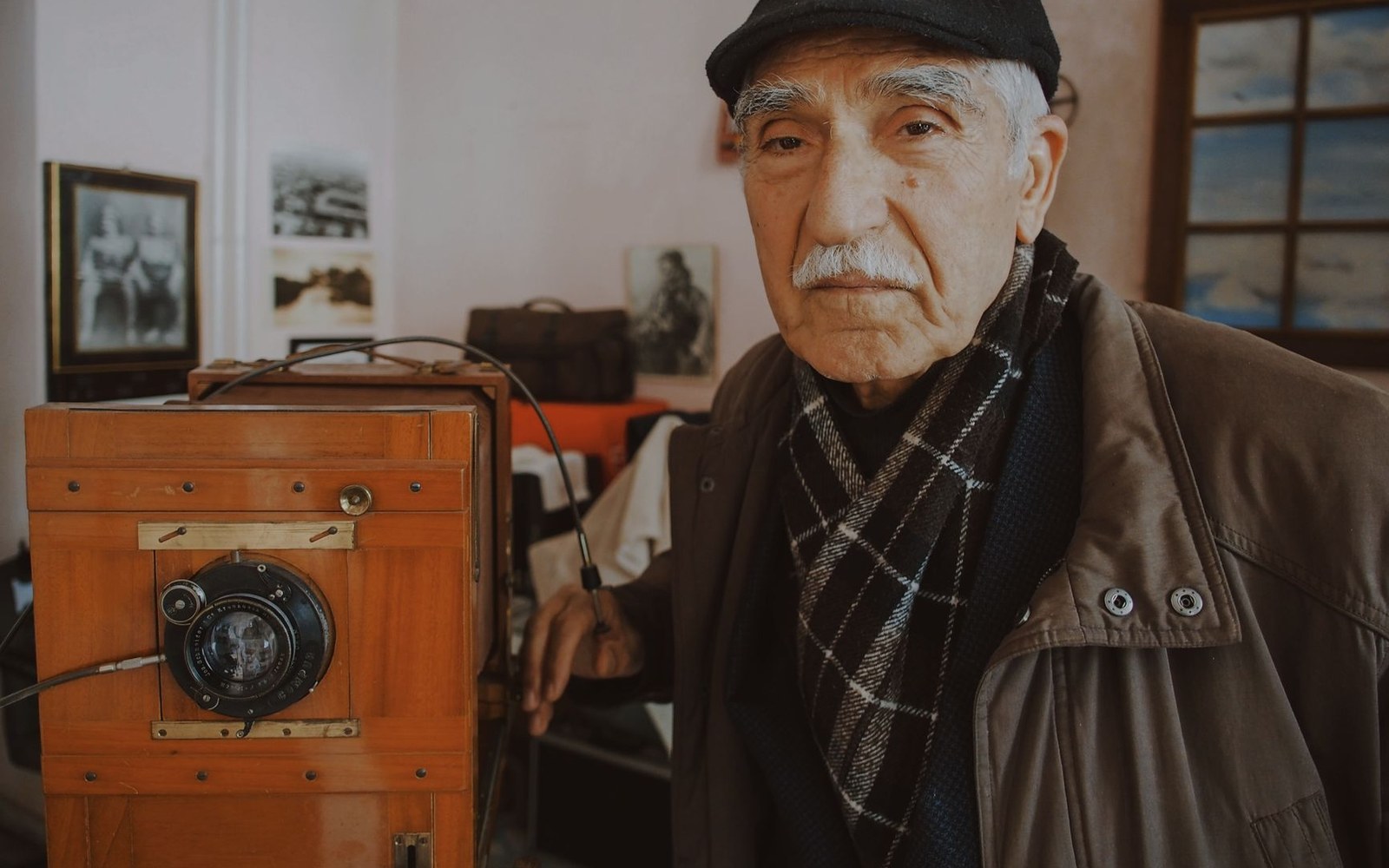 An elderly man in a flat cap and scarf stands beside a vintage wooden plate camera