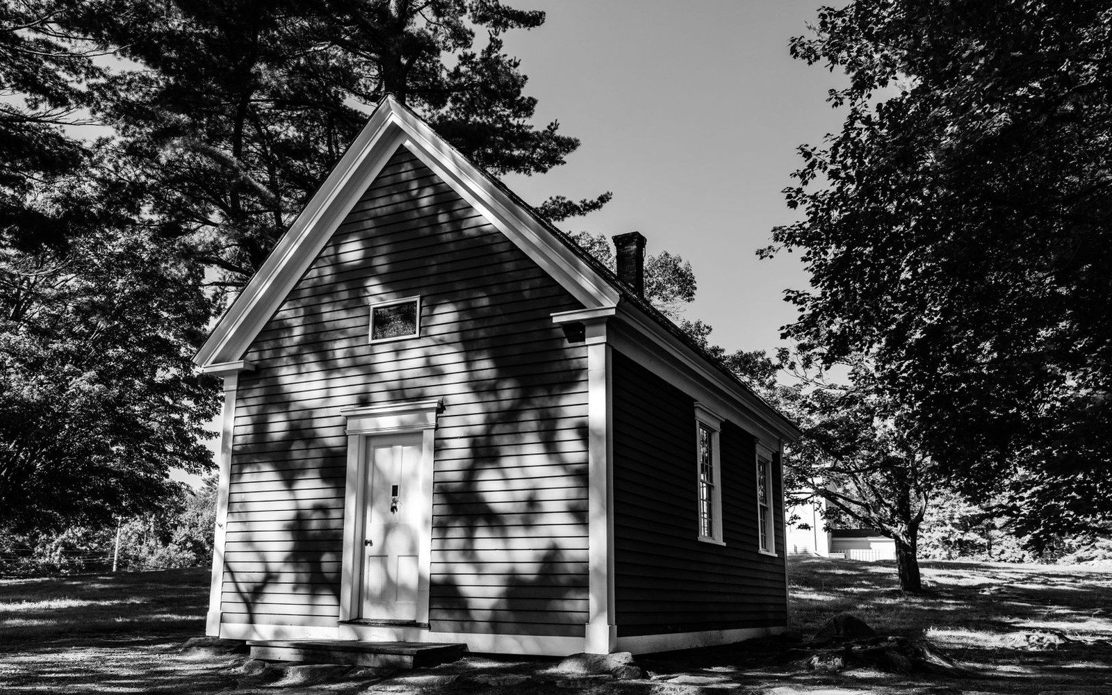 Small clapboard schoolhouse with a peaked roof, shaded by overhanging tree branches