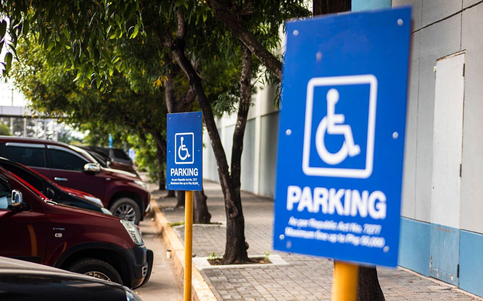 Two blue accessible parking signs line a curbside street with parked vehicles nearby