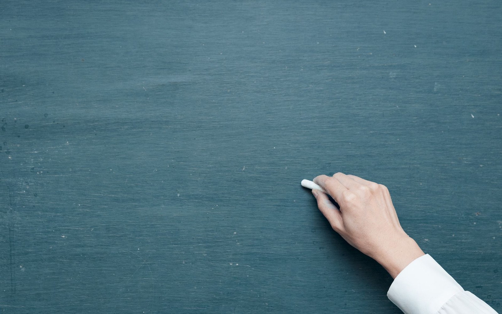 An adult teacher — back partly to camera — writing on a slate chalkboard in an empty classroom, late afternoon light from the windows